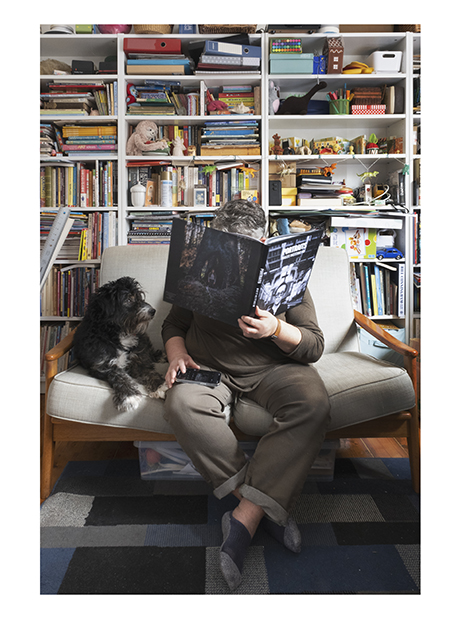 portrait of woman sitting on couch, reading a book, with a dog sitting beside her and looking up at her.